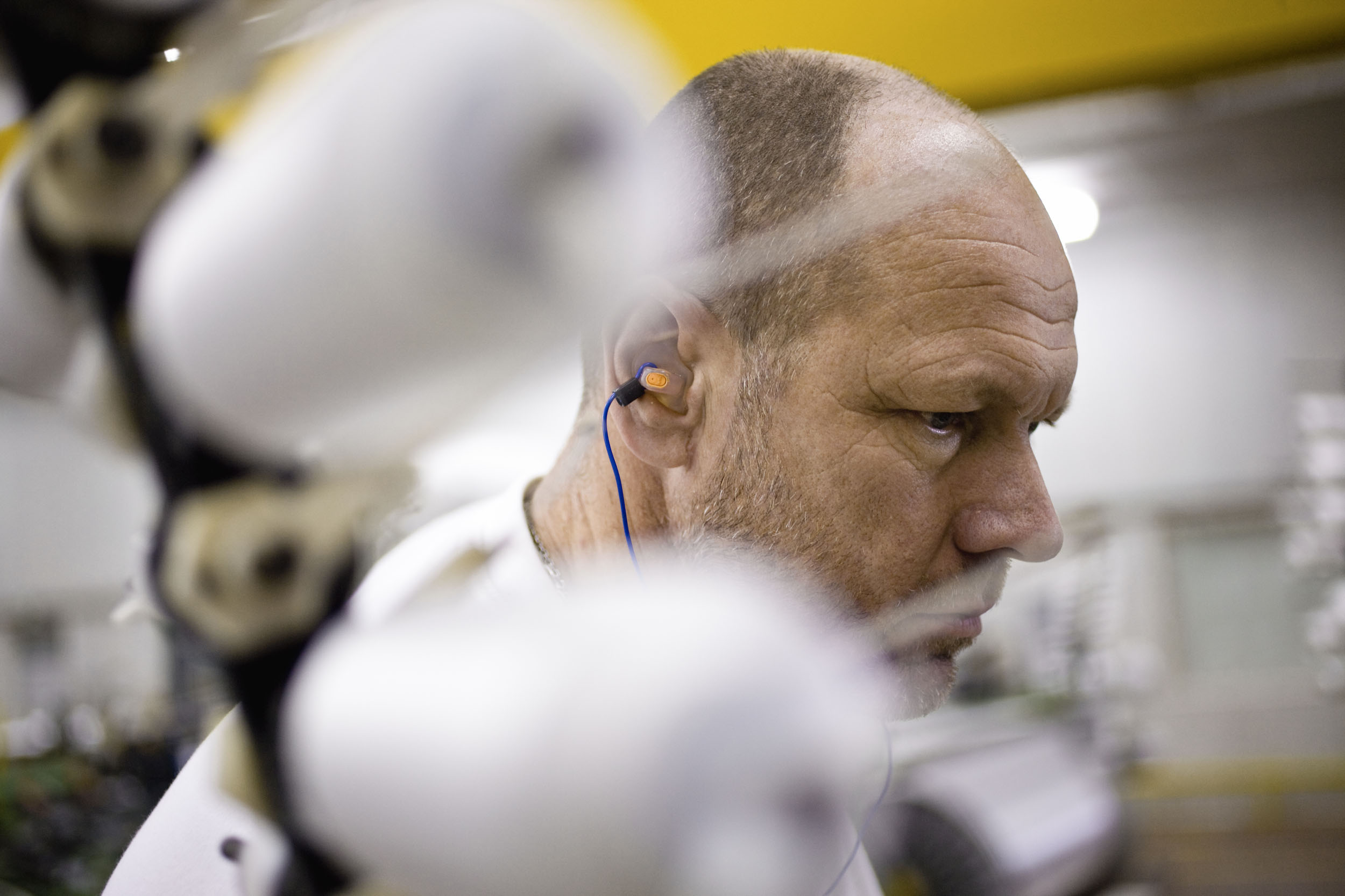 Portrait d'un homme au travail sur une machine dans un atelier de fabrication de textiles, le premier plan est flou. Il porte des bouchons d'oreilles et regarde devant lui vers le bas