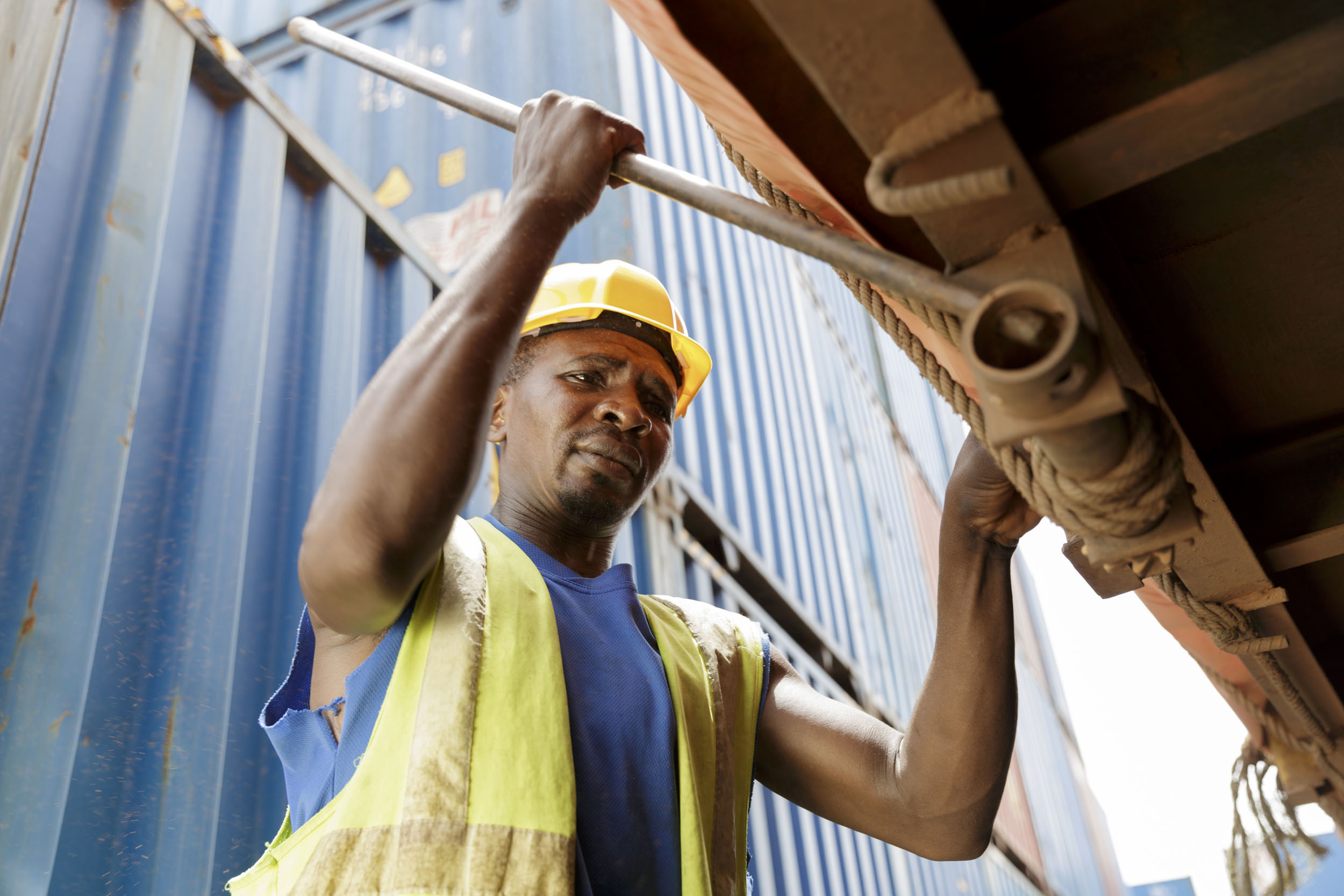 Portrait en contre plongée d'un homme au travail portant un casque et un gilet jaune sur un site industriel de conteneurs au Cameroun, il desserre les attaches d'une bâche de camion