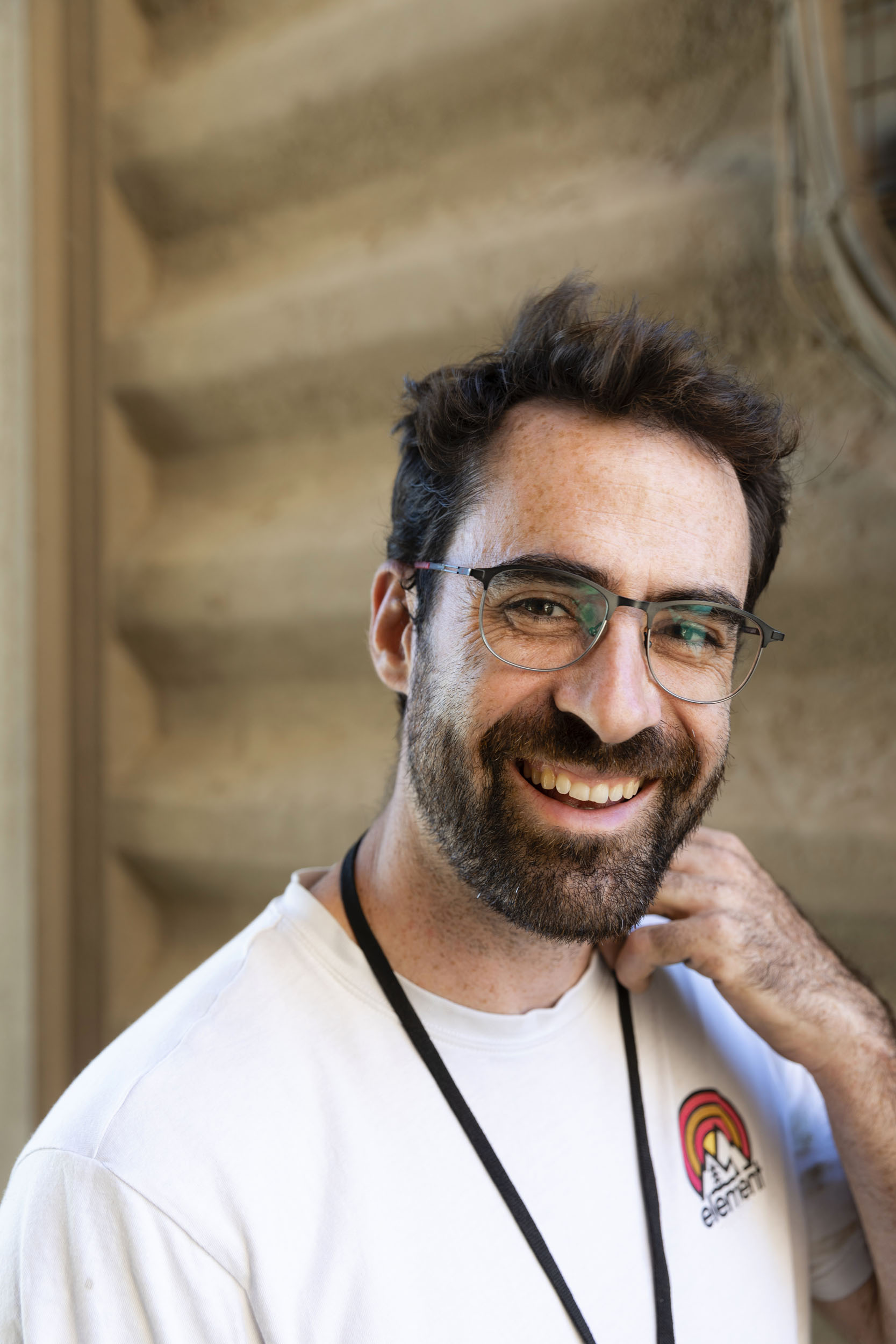 Portrait d'un jeune ingénieur souriant en lumière du jour au pied d'un silo agricole, il est brun, porte un T-shirt blanc, des lunettes, la barbe et la moustache