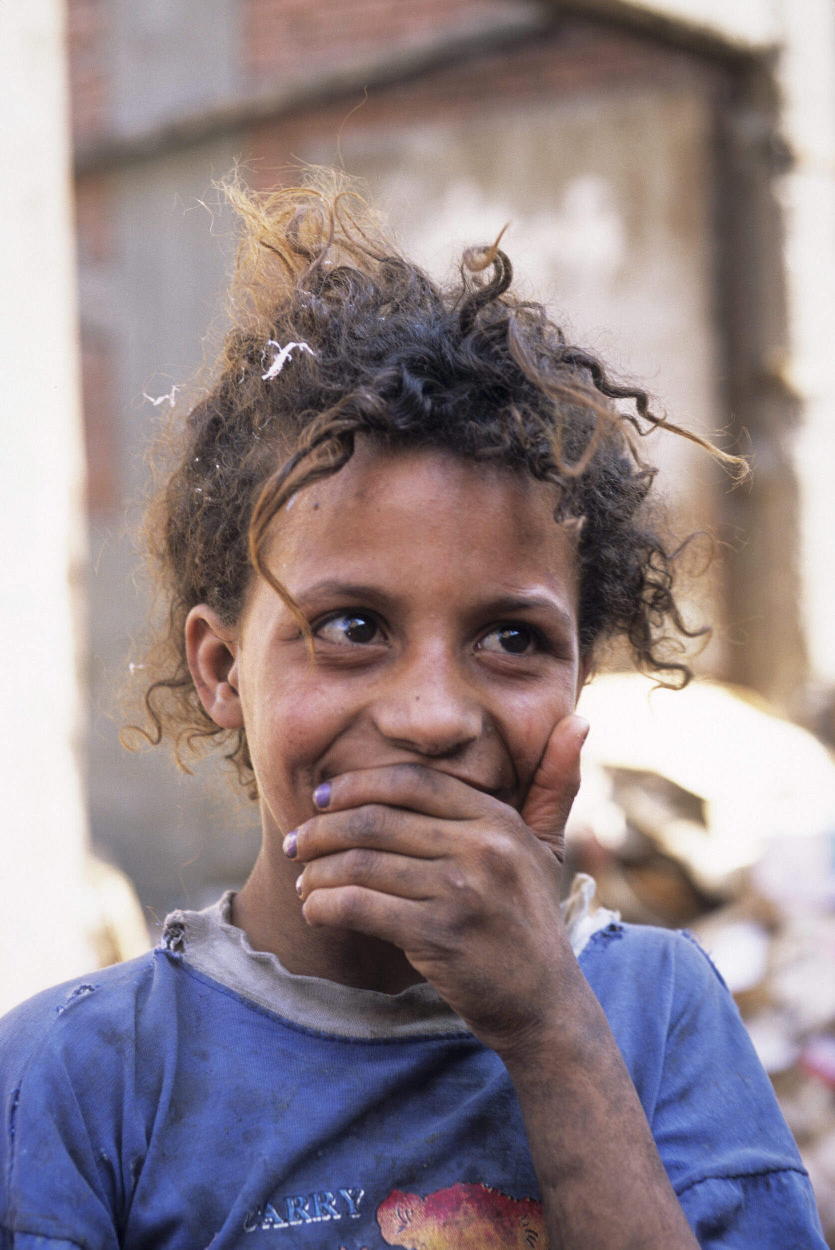 Portrait d'une enfant Zabbaline aux cheveux bouclés et attachés qui tient sa main gauche très sale devant la bouche, elle porte un T-shirt bleu usé