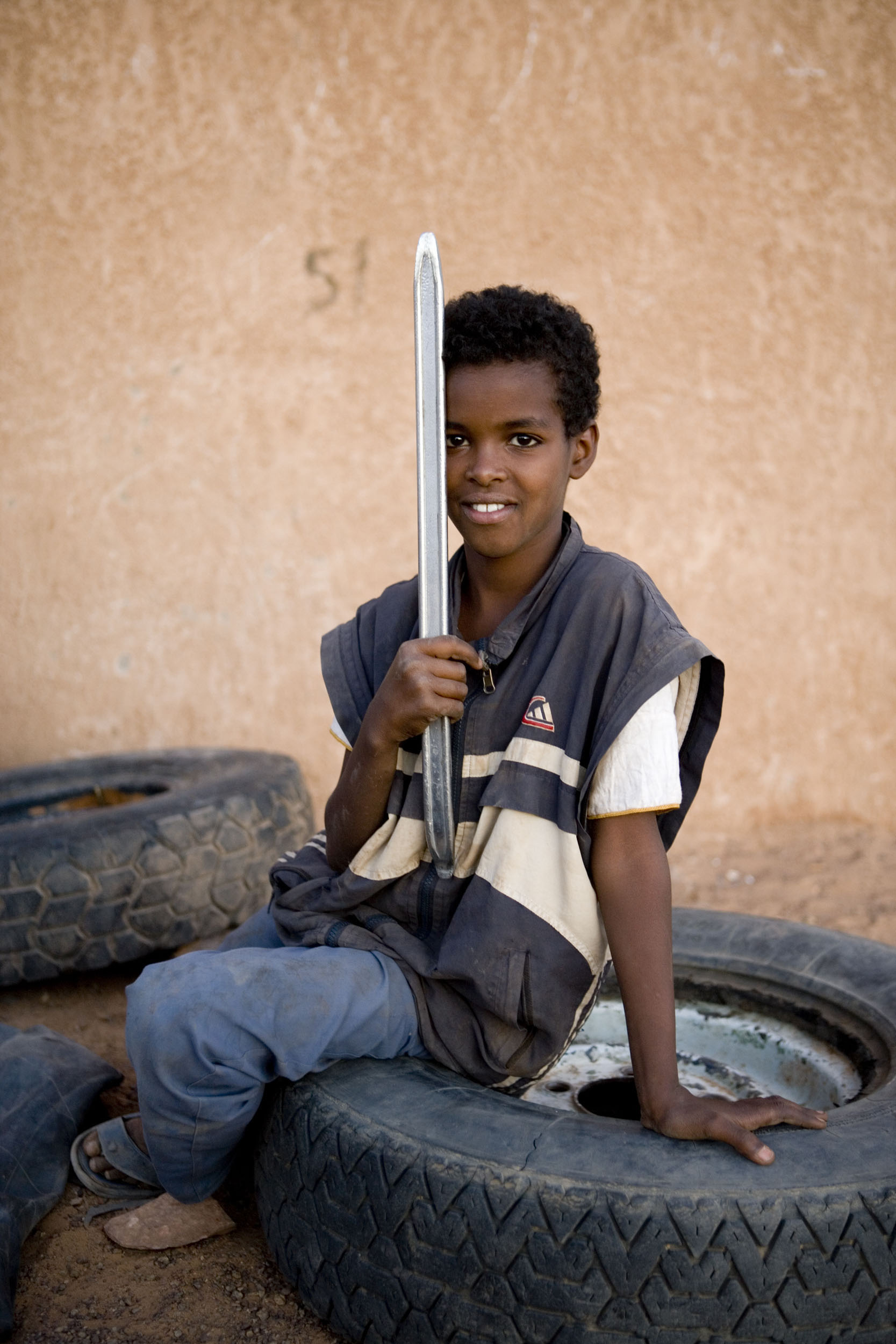 Portrait d'un jeune garçon mauritanien souriant, assis sur un gros pneu de camion dans la rue, un grand démonte pneu à la main, il regarde dans l'objectif