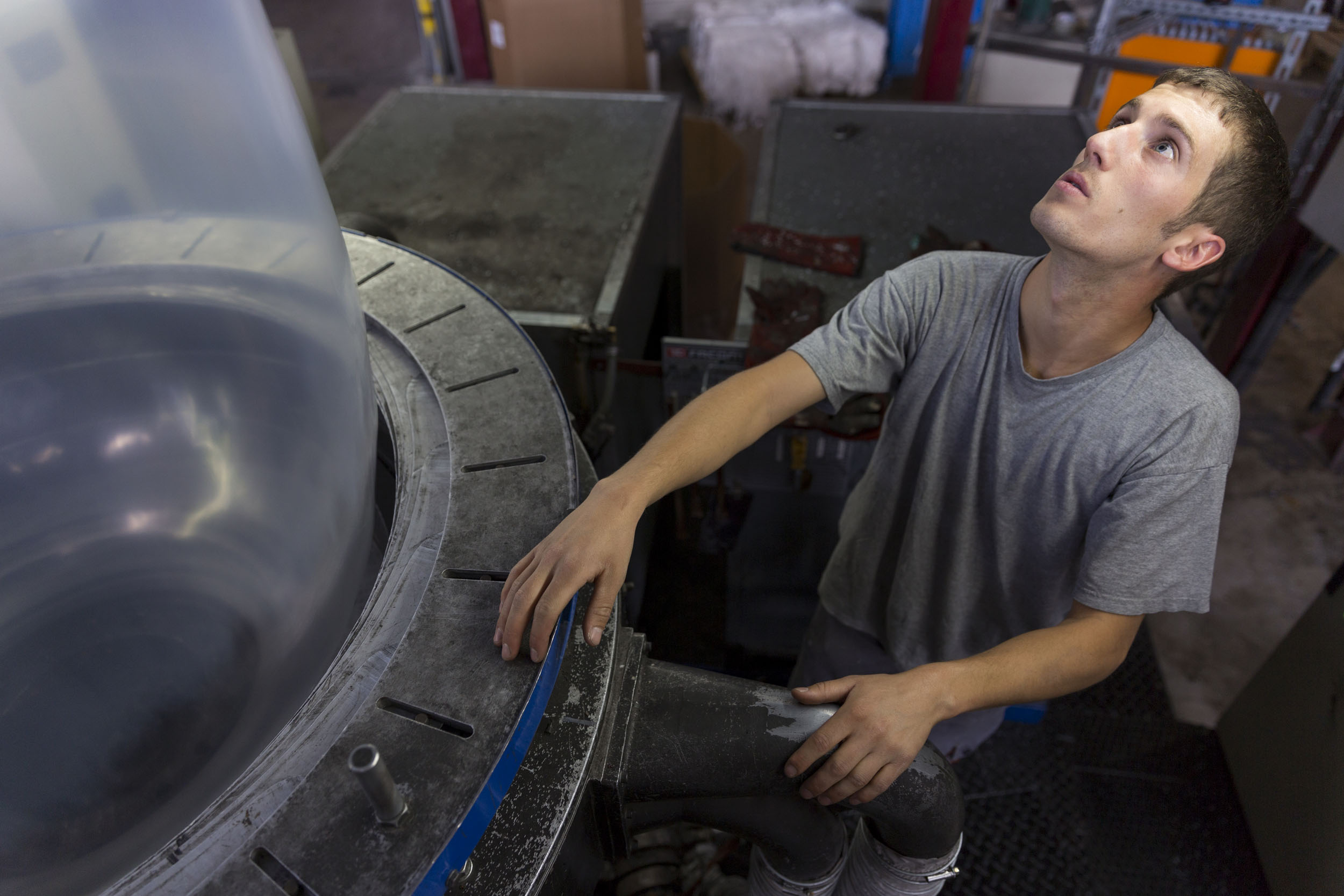 Portrait en plongée d'un jeune ouvrier au travail dans une usine de fabrication de films et membranes techniques. Il porte un T-shirt gris et regarde au-dessus de lui pour surveiller la machine