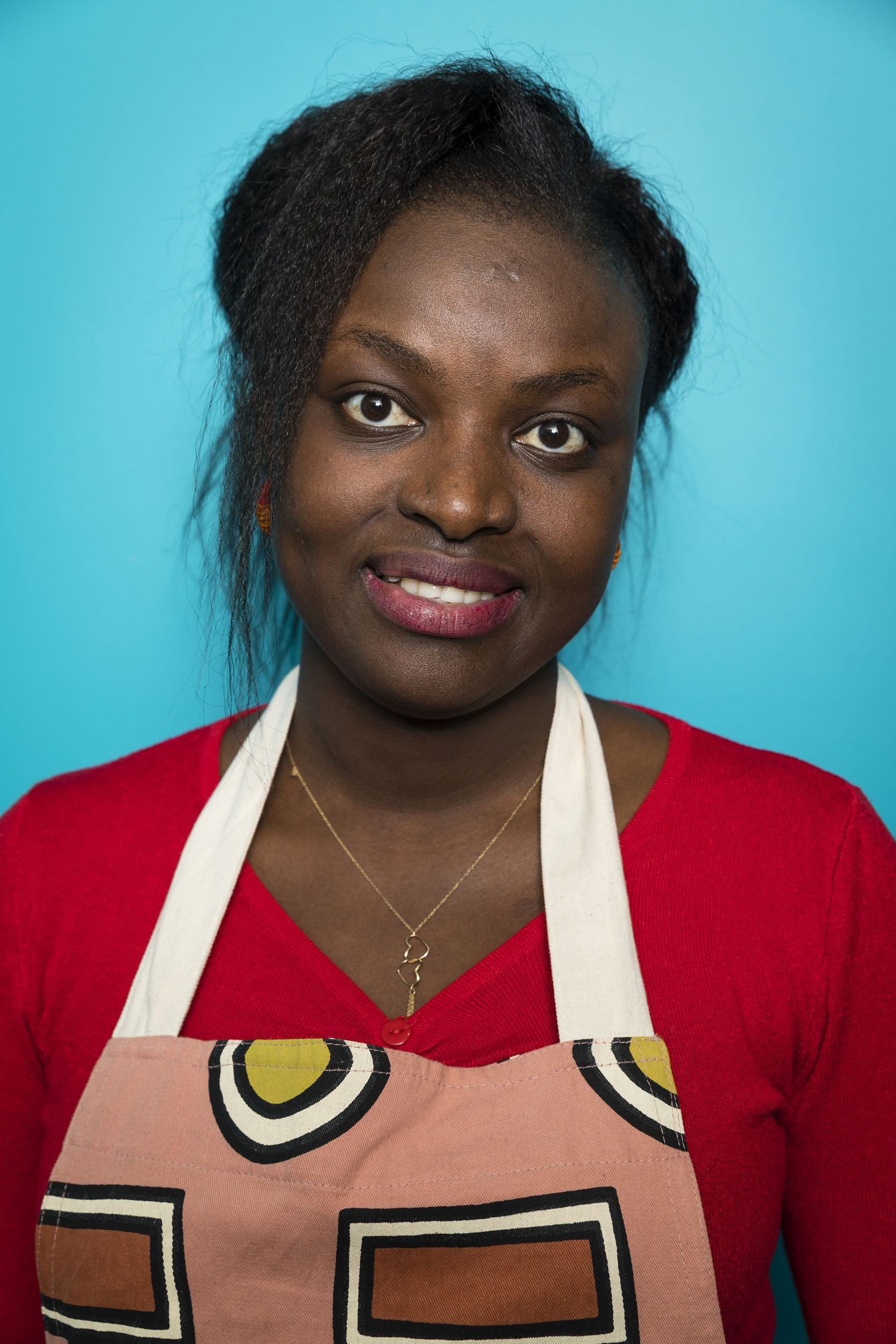 Portrait frontale d'une jeune femme malgache portant un pull rouge et un tablier de cuisine sur fond de mur bleu turquoise