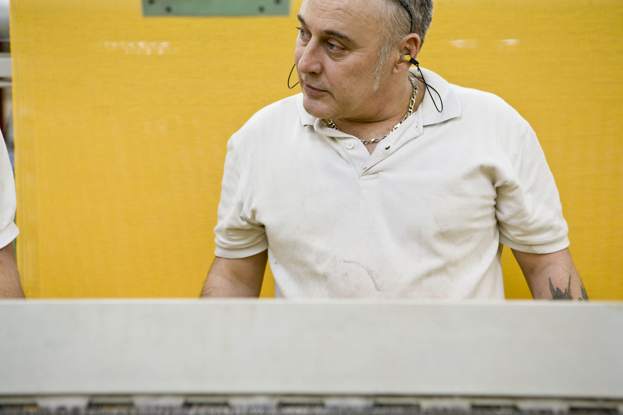Portrait d'un homme portant des bouchons d'oreilles devant un métier à tisser. Il porte un polo blanc et le fond est jaune. Il regarde sur sa droite