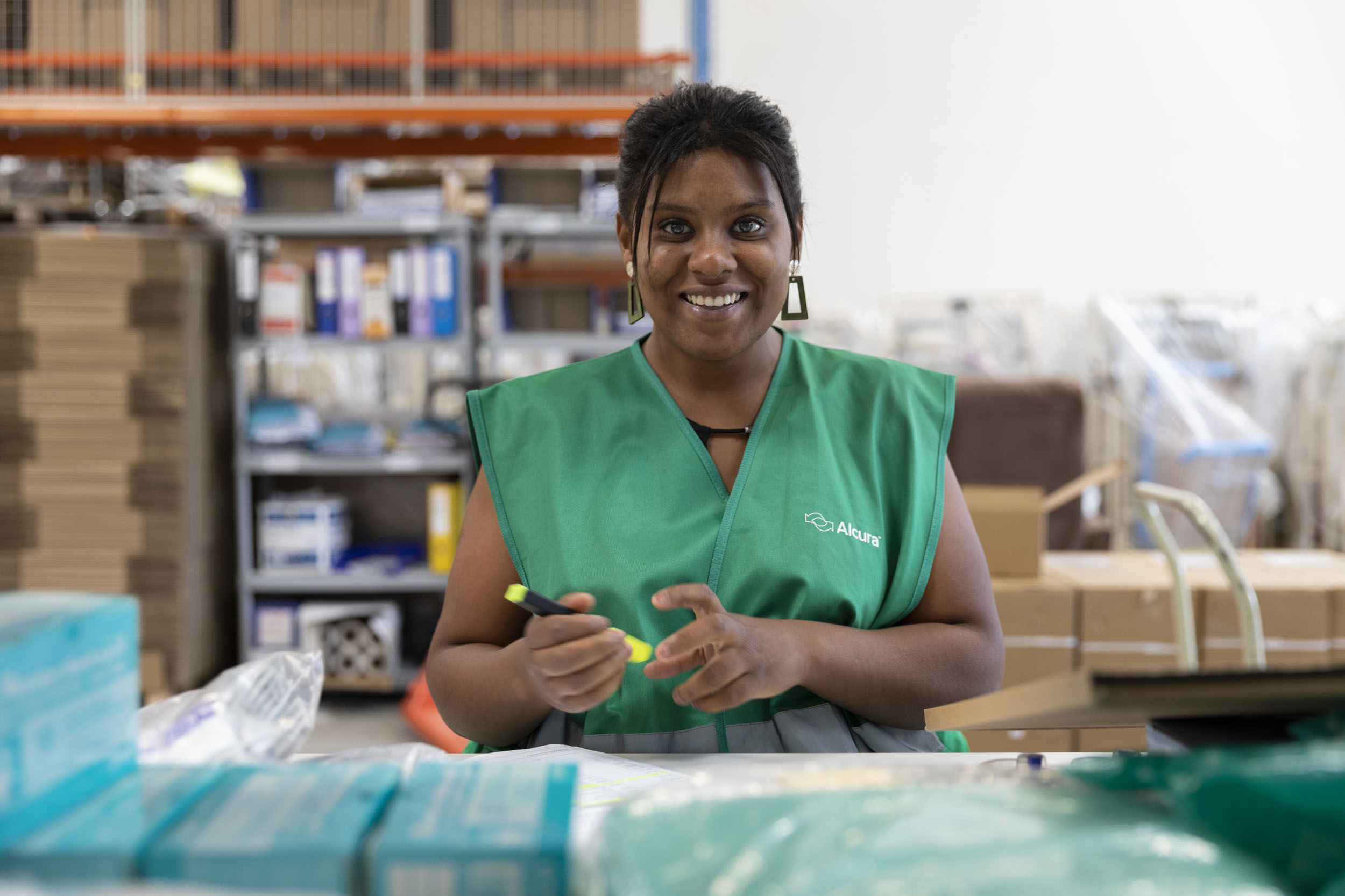 Portrait métier d'une préparatrice de commande dans un entrepôt tenant un surligneur fluorescent jaune dans la main droite. Elle porte un baudrier vert et regarde en souriant l'objectif