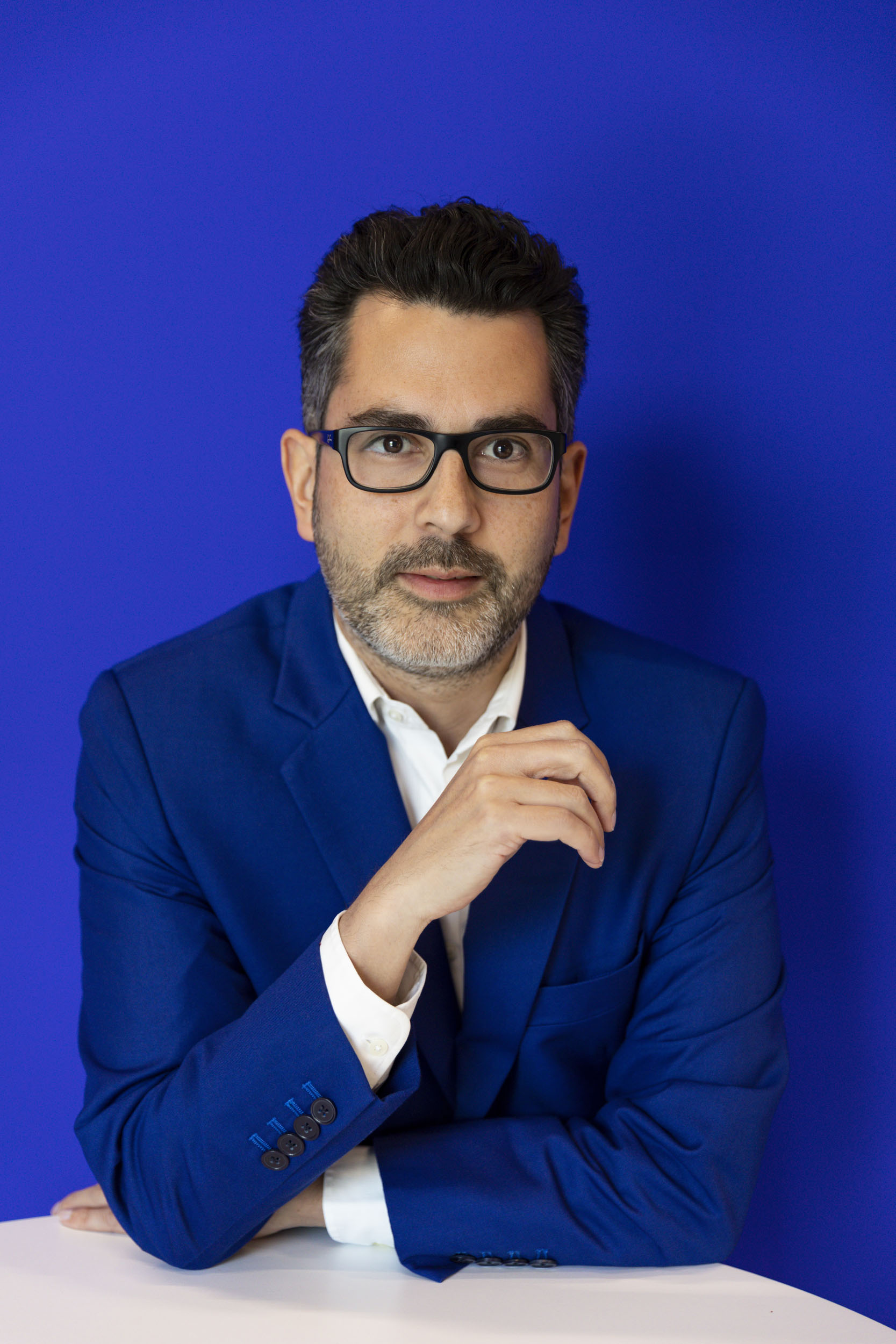 Portrait de Maître Alexandre Poujade, Notaire, réalisée en lumière du jour sur fond bleu. Il est accoudé à un mange debout, porte une veste bleue et regarde dans l'objectif