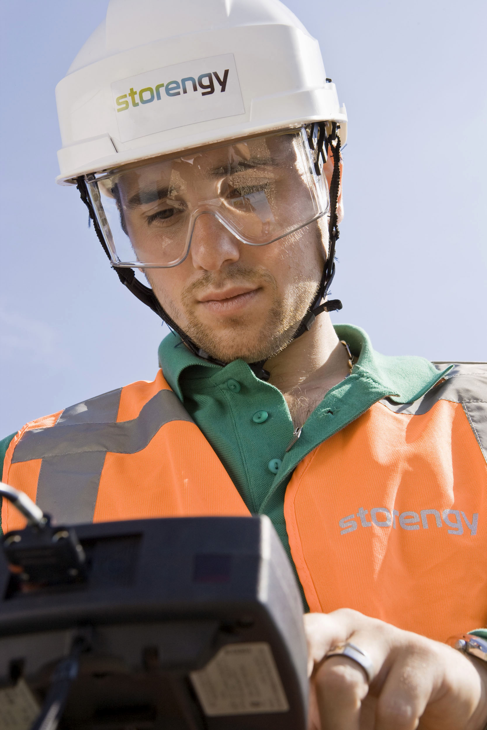 Portrait en contre-plongée d'un homme au travail sur un stockage souterrain de gaz naturel qui porte un casque blanc et des lunettes de protection, il contrôle un capteur de pression à l'aide d'un appareil de mesure