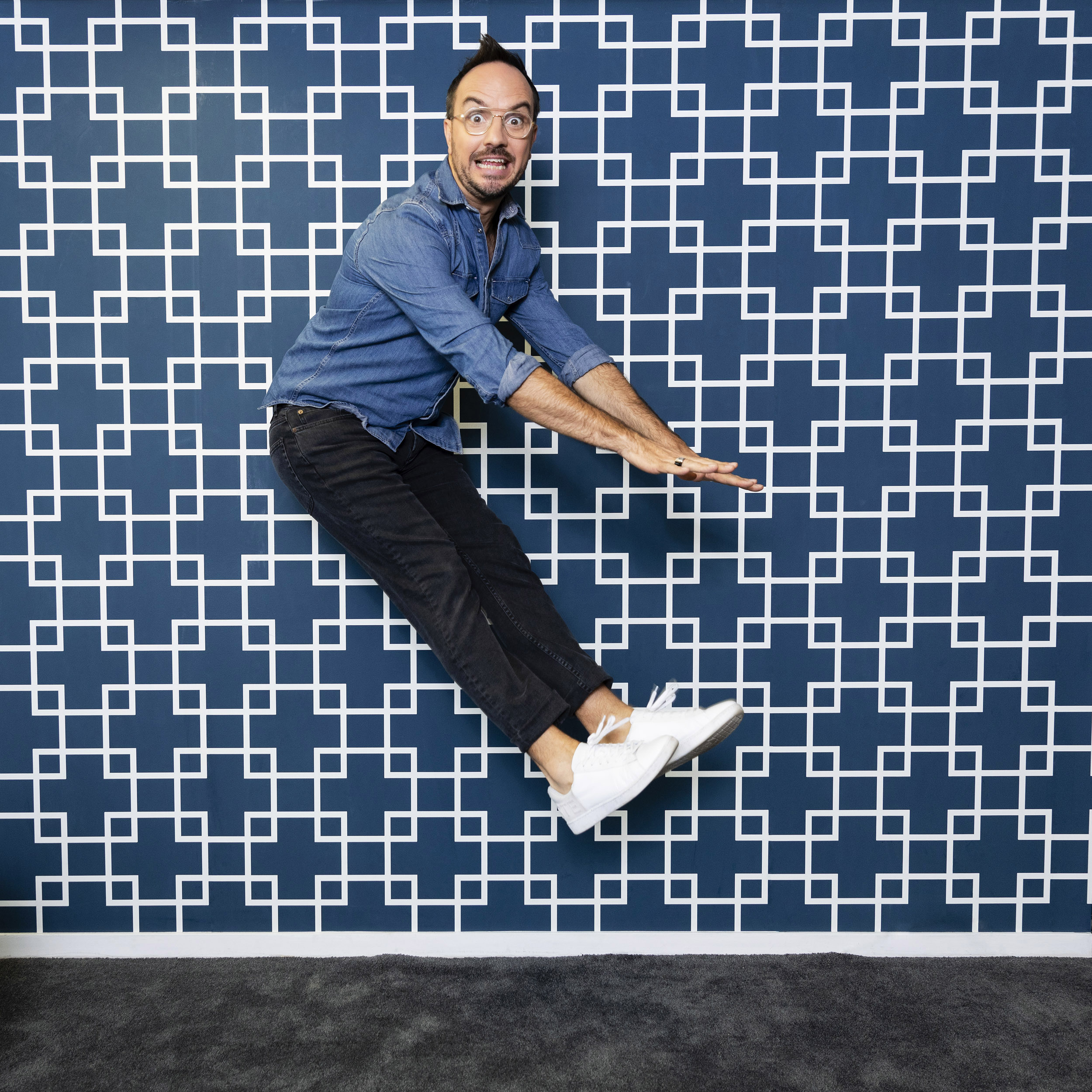 Portrait en pied de l'animateur Jarry qui saute en l'air en regardant dans l'objectif, il porte une chemise en jean bleu, un jean noir et des sneakers blanches. Le mur est couvert de papier peint bleu avec des motifs blancs