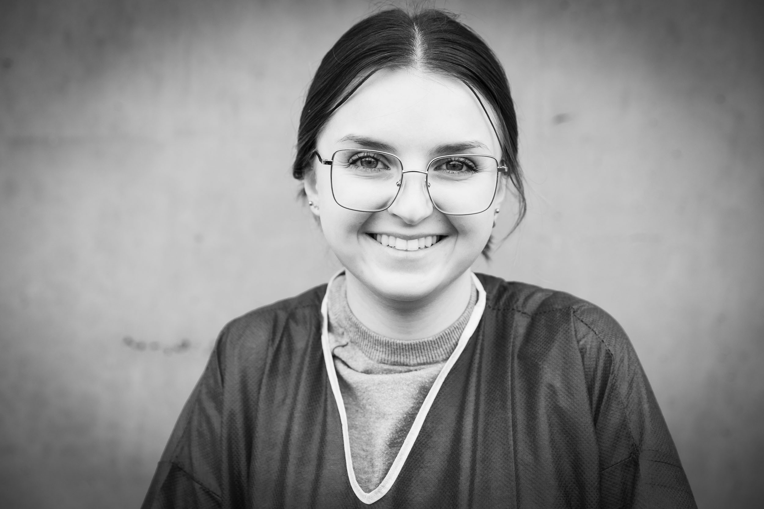 Portrait noir et blanc en lumière du jour d'une jeune soignante à l'hôpital, elle est souriante, porte des lunettes et regarde dans l'objectif