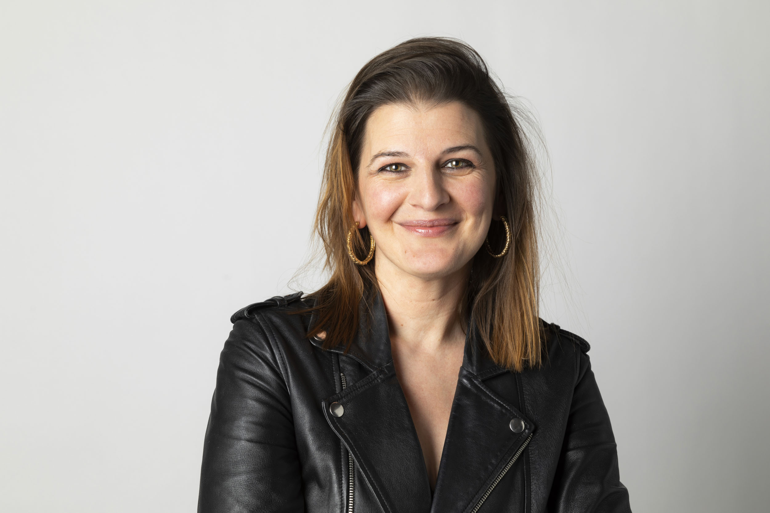 Portrait corporate souriant d'une femme en studio sur fond blanc cadré en plan buste. Elle porte un blouson noir Perfecto et des boucles d'oreilles créoles