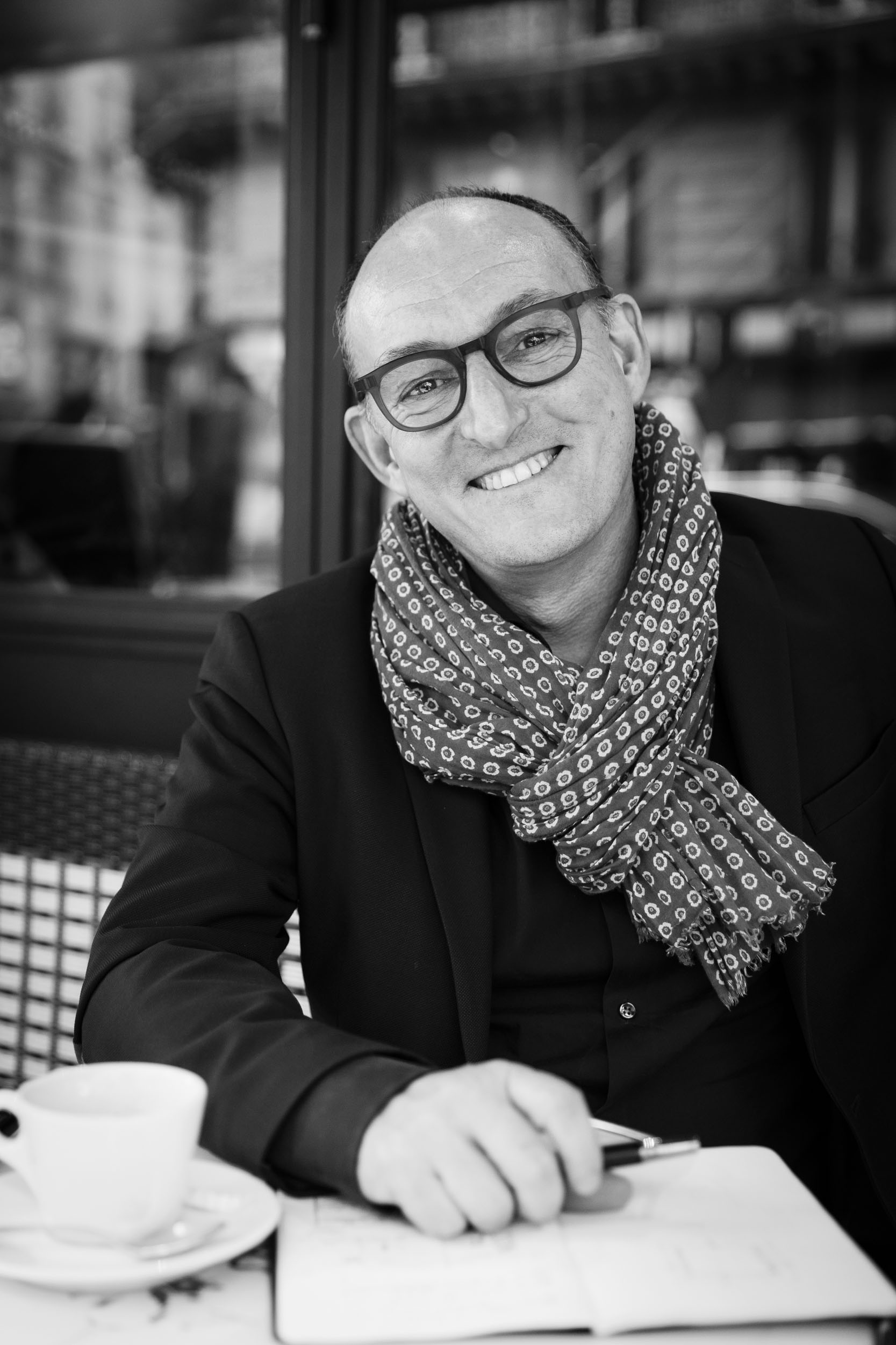 Portrait en noir et blanc de l'architecte d'intérieur Yann Montfort souriant assis à la terrasse d'un café parisien, il tient un crayon à la main, son carnet de croquis est posé sur la table