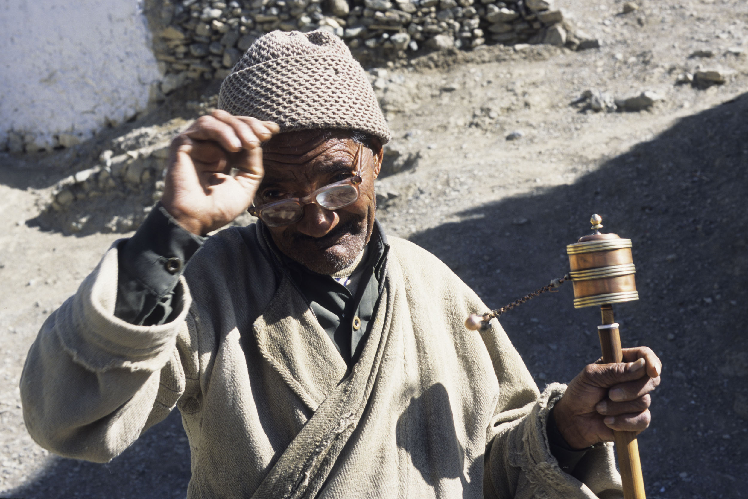 Portrait argentique d'un vieil homme portant un bonnet et des lunettes dans la rue au Ladakh en Inde, il fait tourner un moulin à prières avec sa main gauche