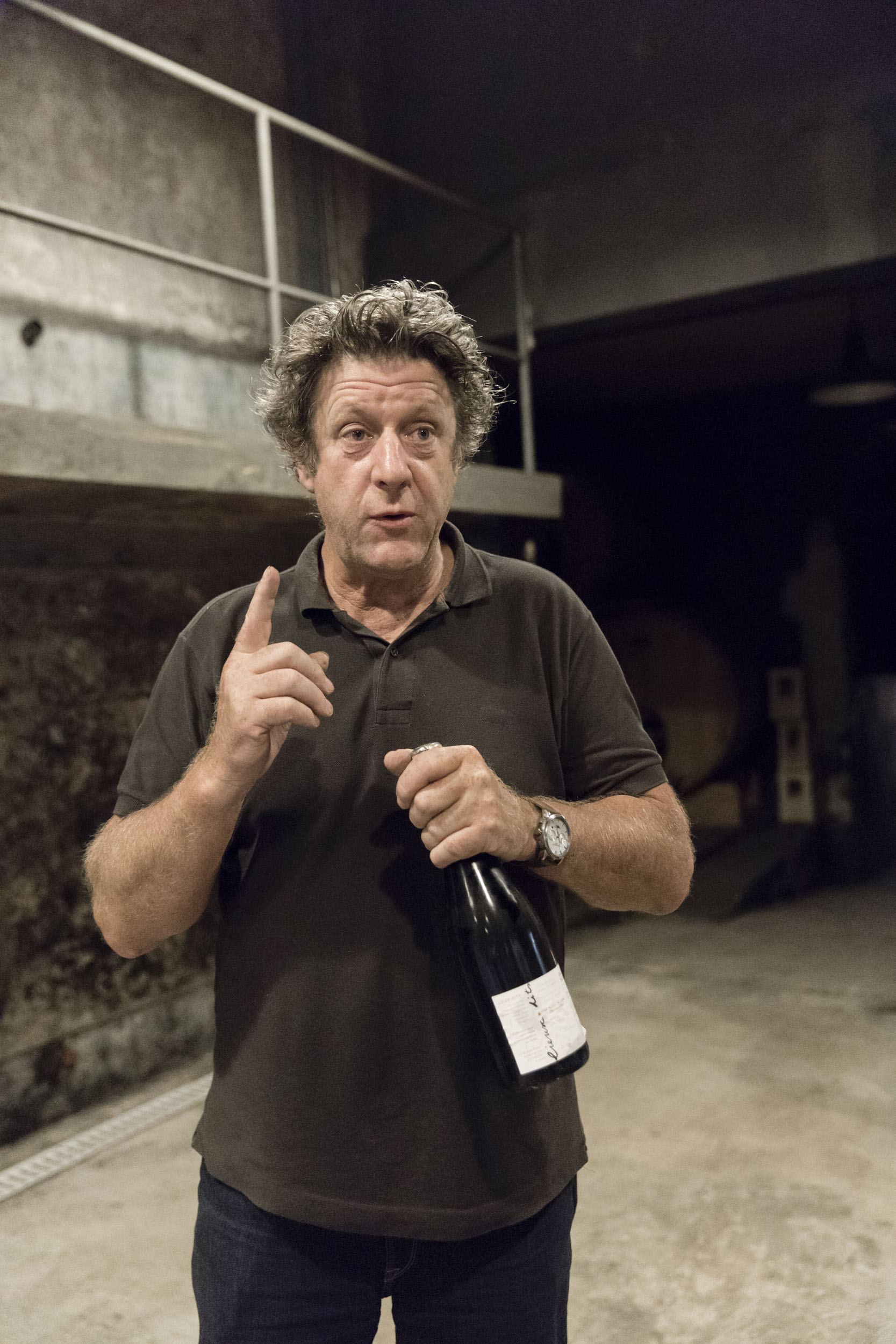 Portrait du vigneron Jacques Selosse dans ses caves, une bouteille de champagne à la main lors d'une dégustation, il porte un polo marron et a l'index de la main droite levé