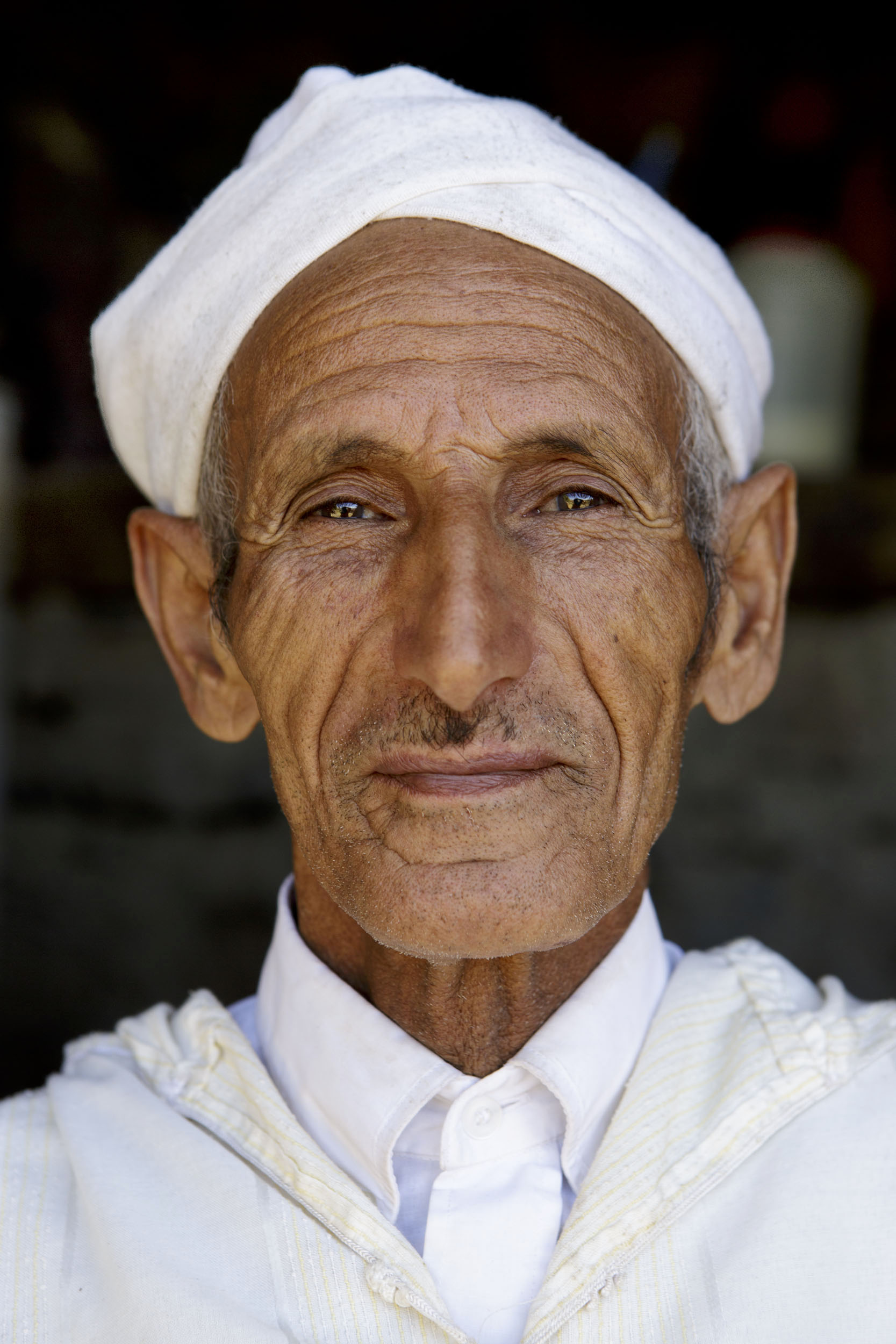 Portrait frontal du visage d'un homme fier en chemise blanche, djellaba et chèche blanc sur la tête, il pose assis devant sa maison dans la vallée d'Anergui au Maroc