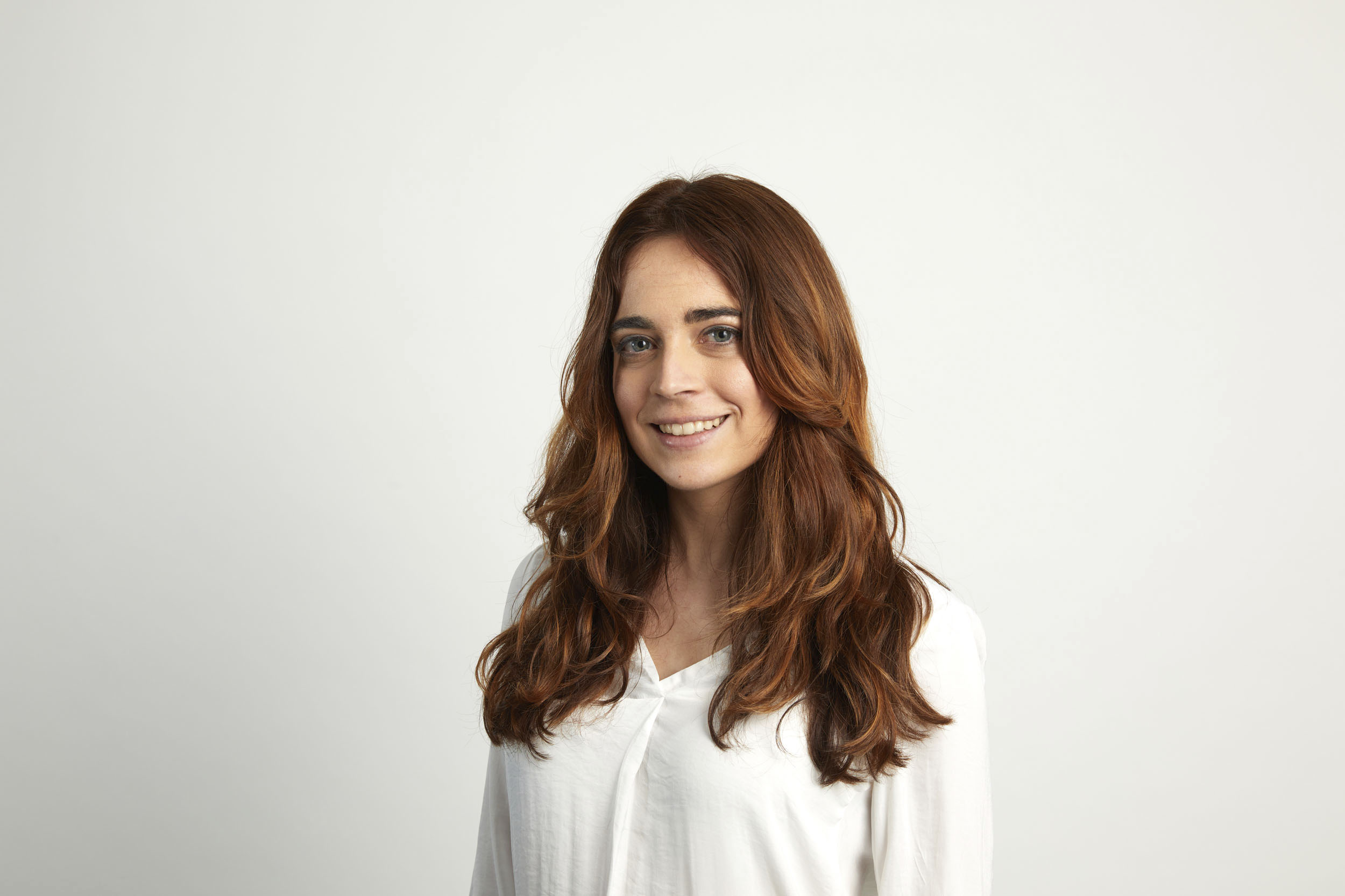 Portrait corporate d'une femme souriante aux cheveux longs vêtue d'un chemisier blanc. Portrait réalisé en studio mobile sur fond blanc et cadrée en plan buste