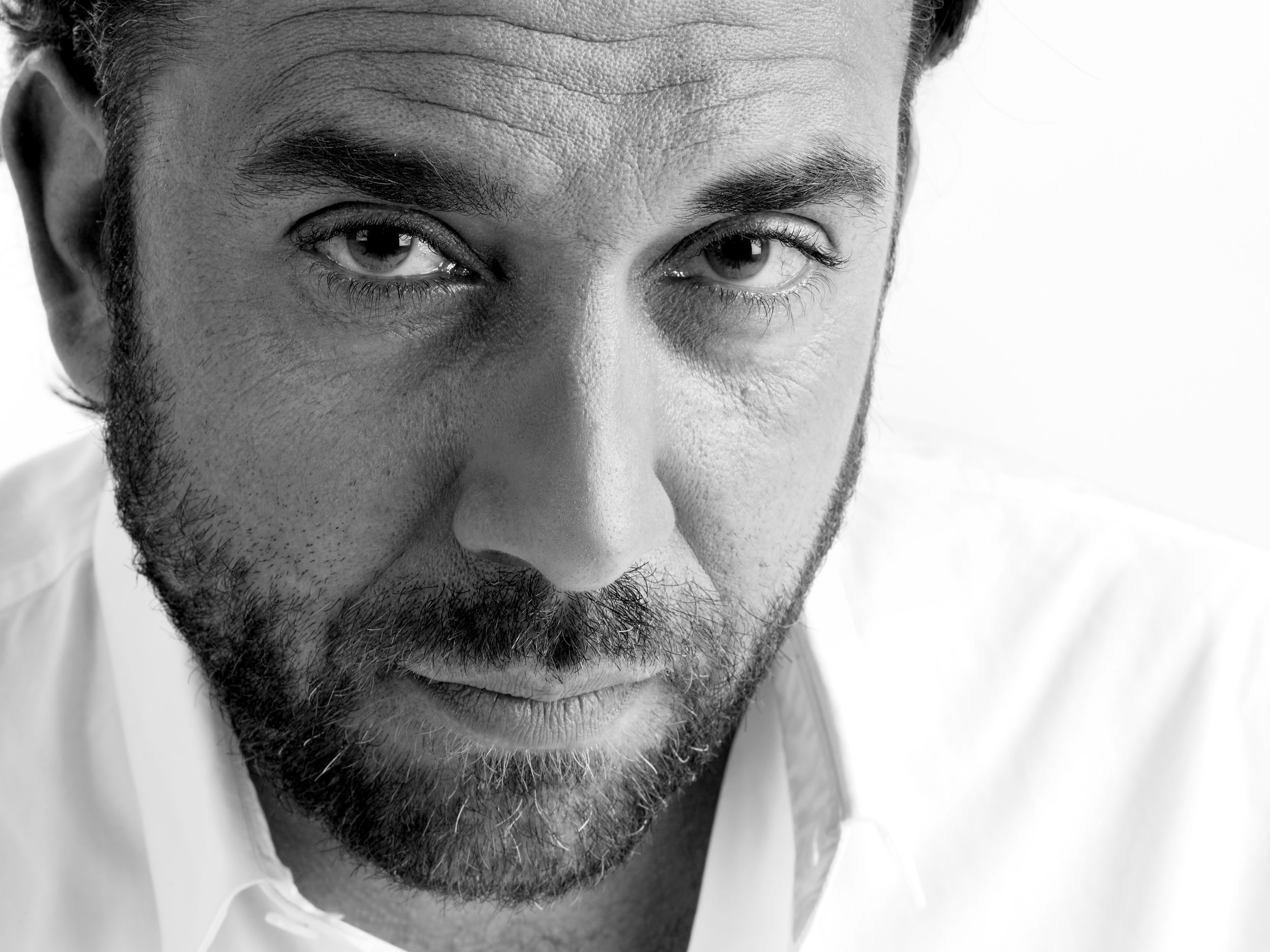 Portrait d'un homme brun portant la barbe et la moustache, en noir et blanc et en gros plan. Il a la tête légèrement penchée. Portrait réalisé en studio sur fond blanc