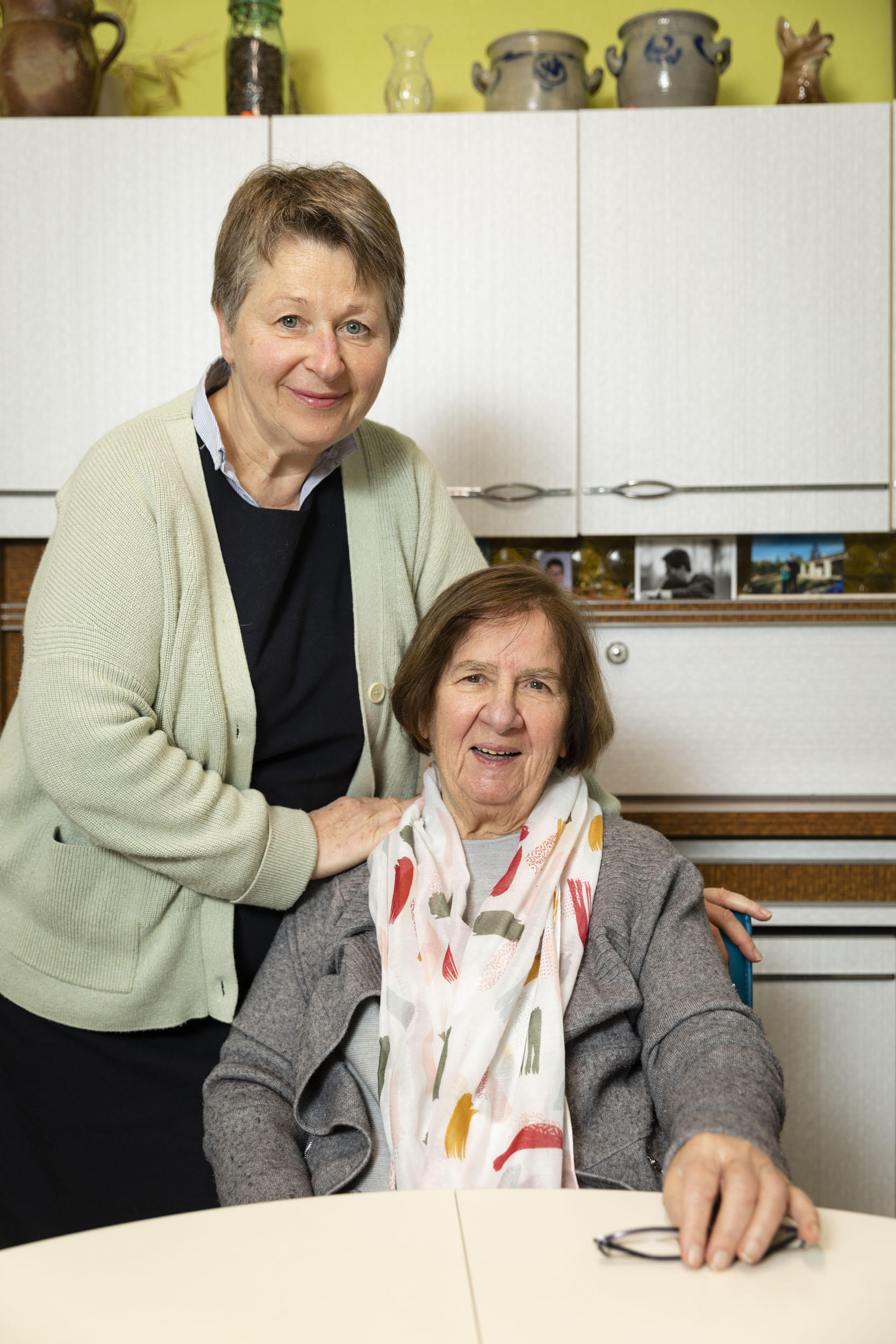 Portrait d'une mère âgée atteinte de la maladie d'Alzheimer assise aux côtés de sa fille qui veille sur elle debout à la cuisine. Elles sourient et regarde dans l'objectif du photographe