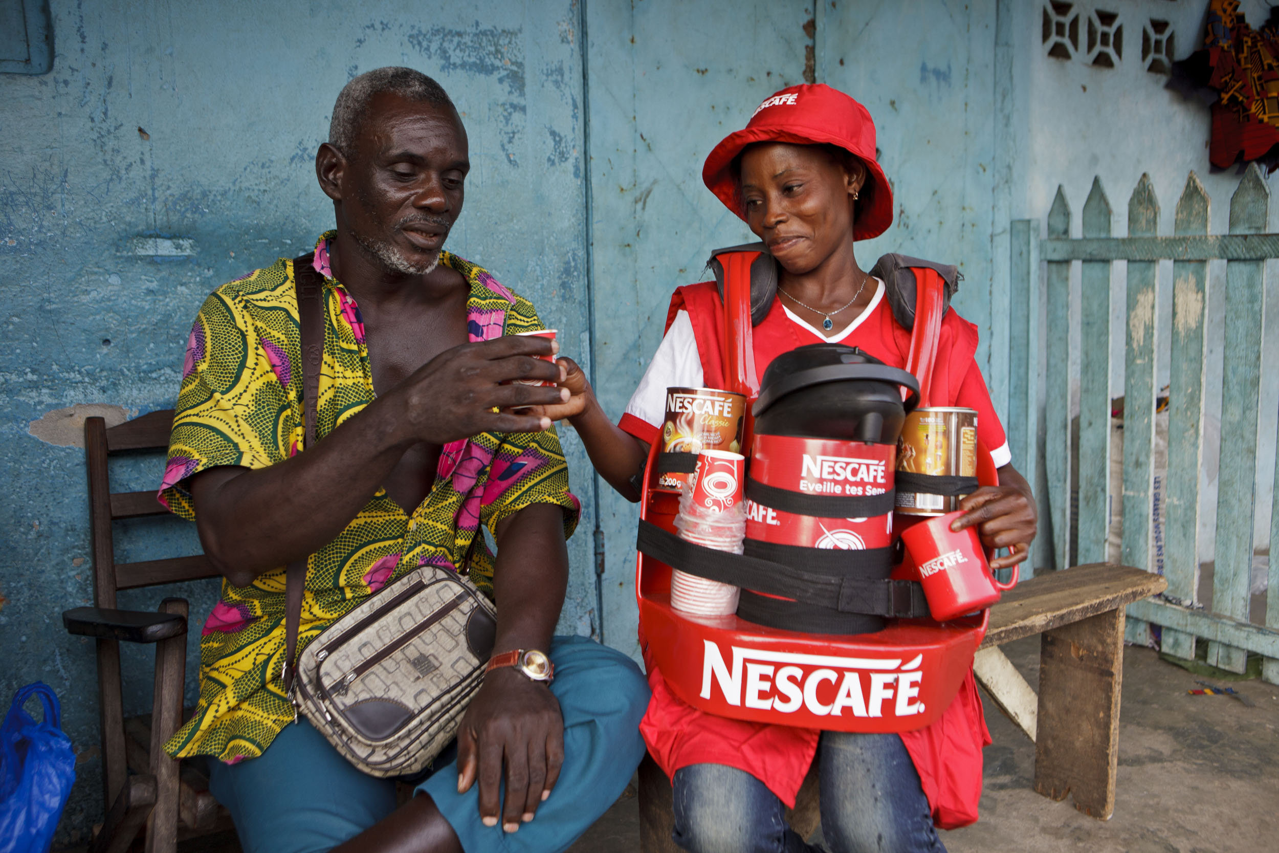 Portrait d'une marchande ambulante de Nescafé® assise sur un banc en train de servir un café à son client assis sur un fauteuil en bois sur un marché en Côte d'Ivoire, le fond est bleu turquoise