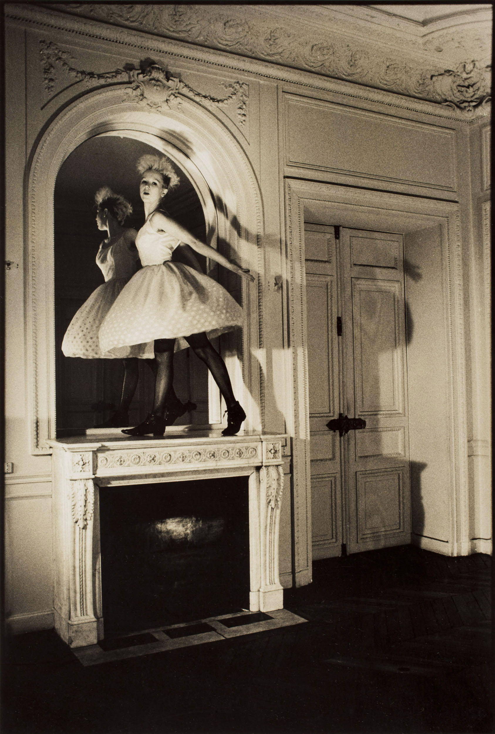 Danseuse debout sur une cheminée en marbre surmontée d'un miroir dans un appartement Haussmannien, photo noir et blanc en plan large