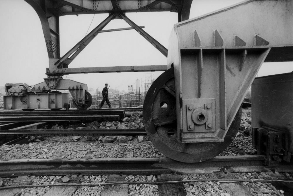 Photo en noir et blanc, roues métalliques d'une grosse grue sur rail au premier plan et un personnage qui marche avec un casque de chantier sur la tête à l'arrière plan