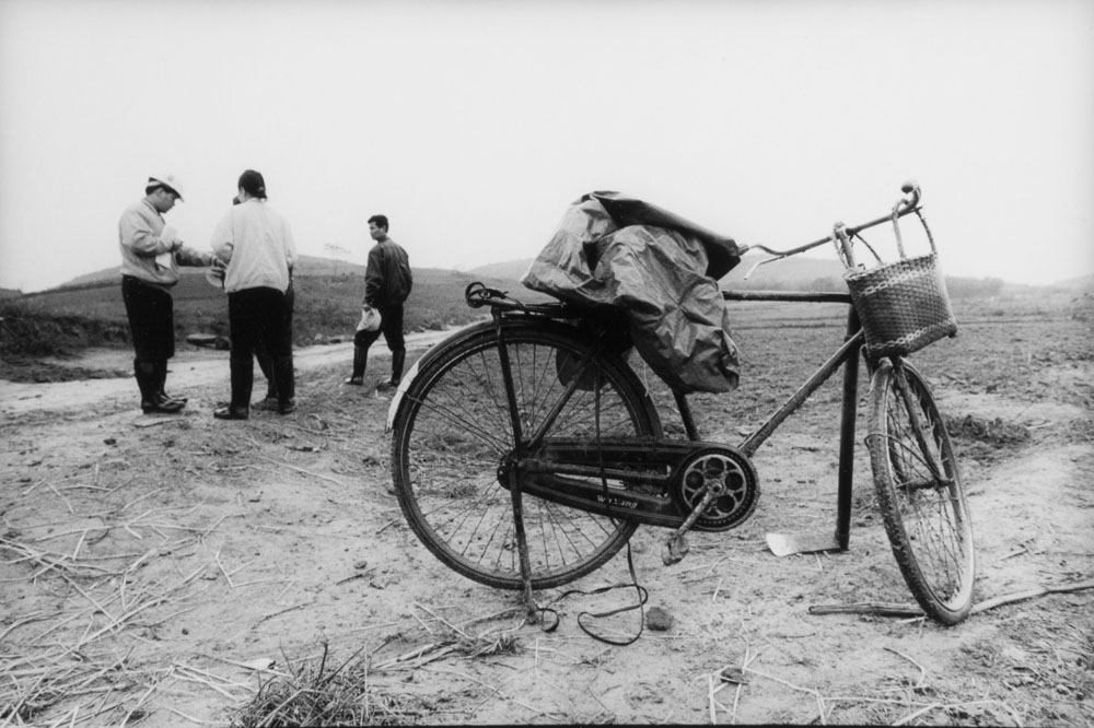 Un vélo dans la campagne chinoise au premier plan avec 4personnages en conversation à l'arrière plan, photo noir et blanc de reportage