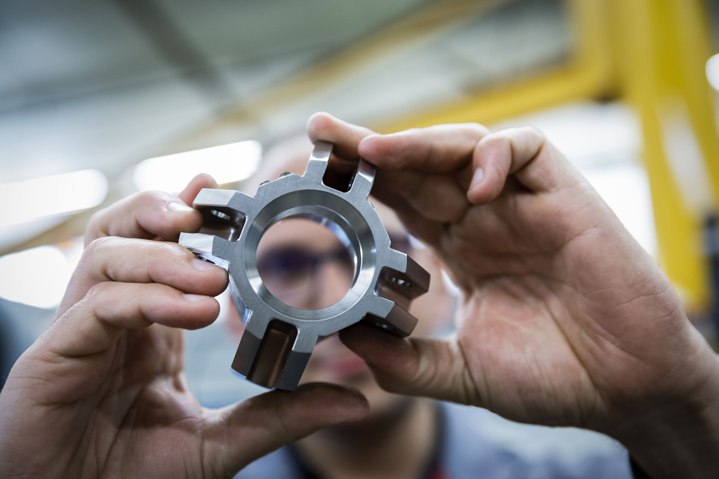 Gros plan en contreplongé sur les 2 mains d'un homme en usine qui réalise un contrôle visuel d'une pièce mécanique. La pièce métallique est net au premier plan, son visage est flou en arrière plan
