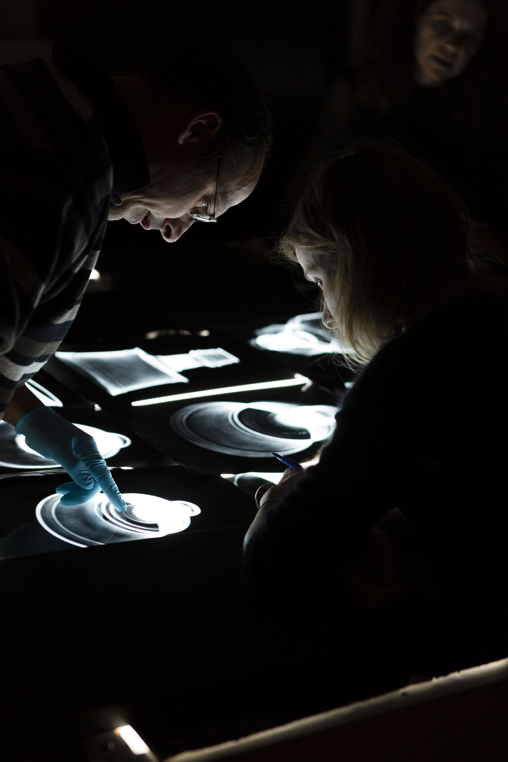 Une chercheuse à droite et un chercheur à gauche analysent des radiographies d'objets d'art posées sur une table lumineuse, la pièce est plongée dans le noir