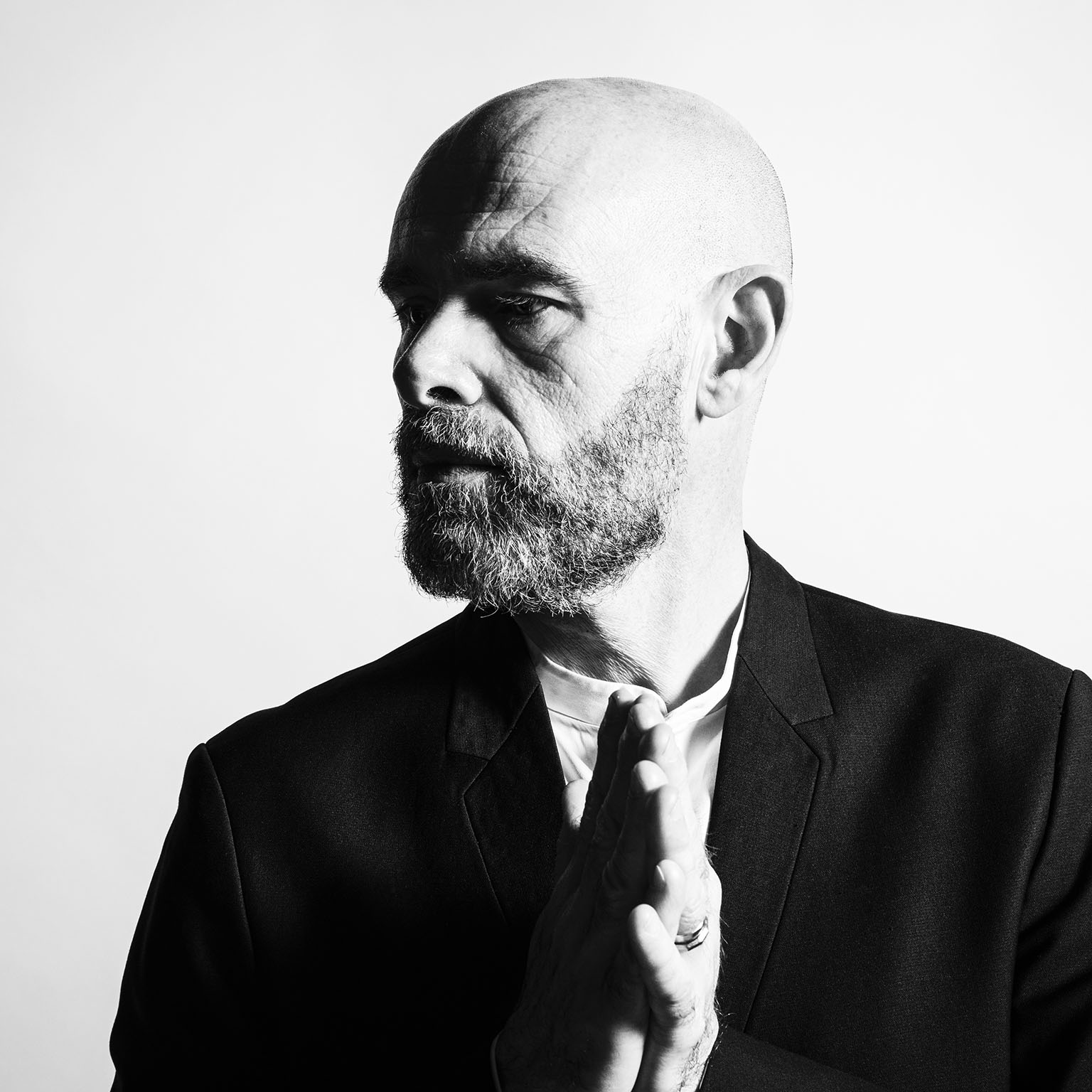 Photo de portrait au format carré d'un homme en noir et blanc très contrasté, il est chauve, barbu, a les mains jointes et regarde sur sa droite
