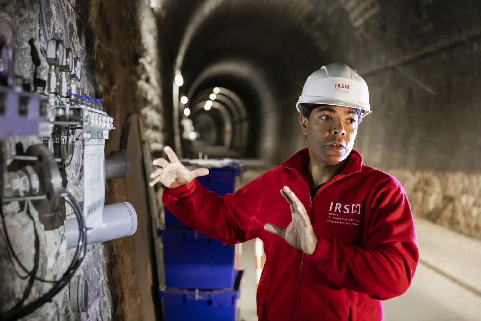 Portrait d'un ingénieur de l'IRSN portant un casque blanc et une veste en polaire rouge dans un tunnel souterrain, il donne des explications à de visiteurs