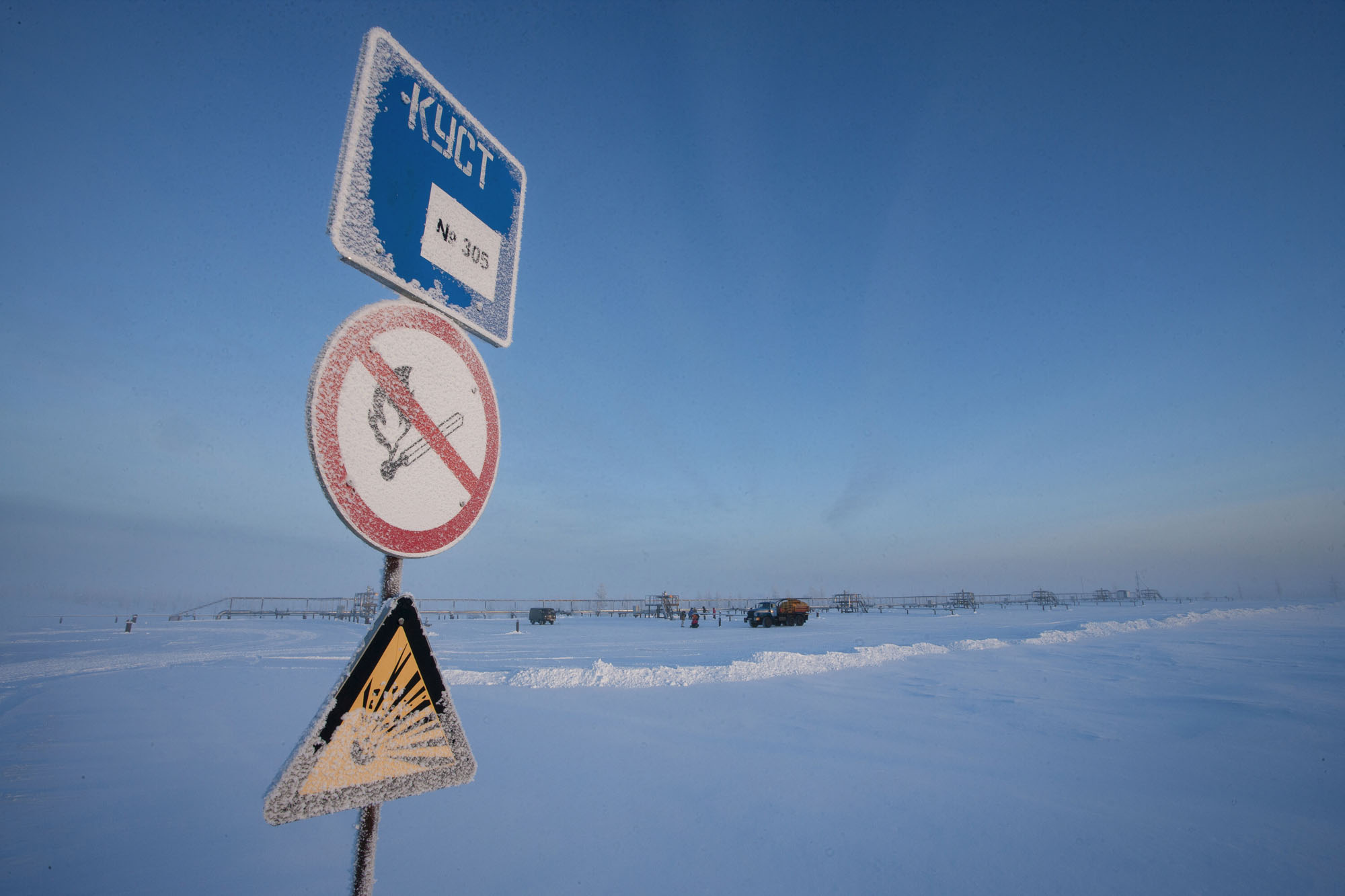 Stockage de gaz en Sibérie en hiver, le sol est couvert de neige, le ciel est bleu, 3 panneaux de signalisation givrés sont visibles au premier plan