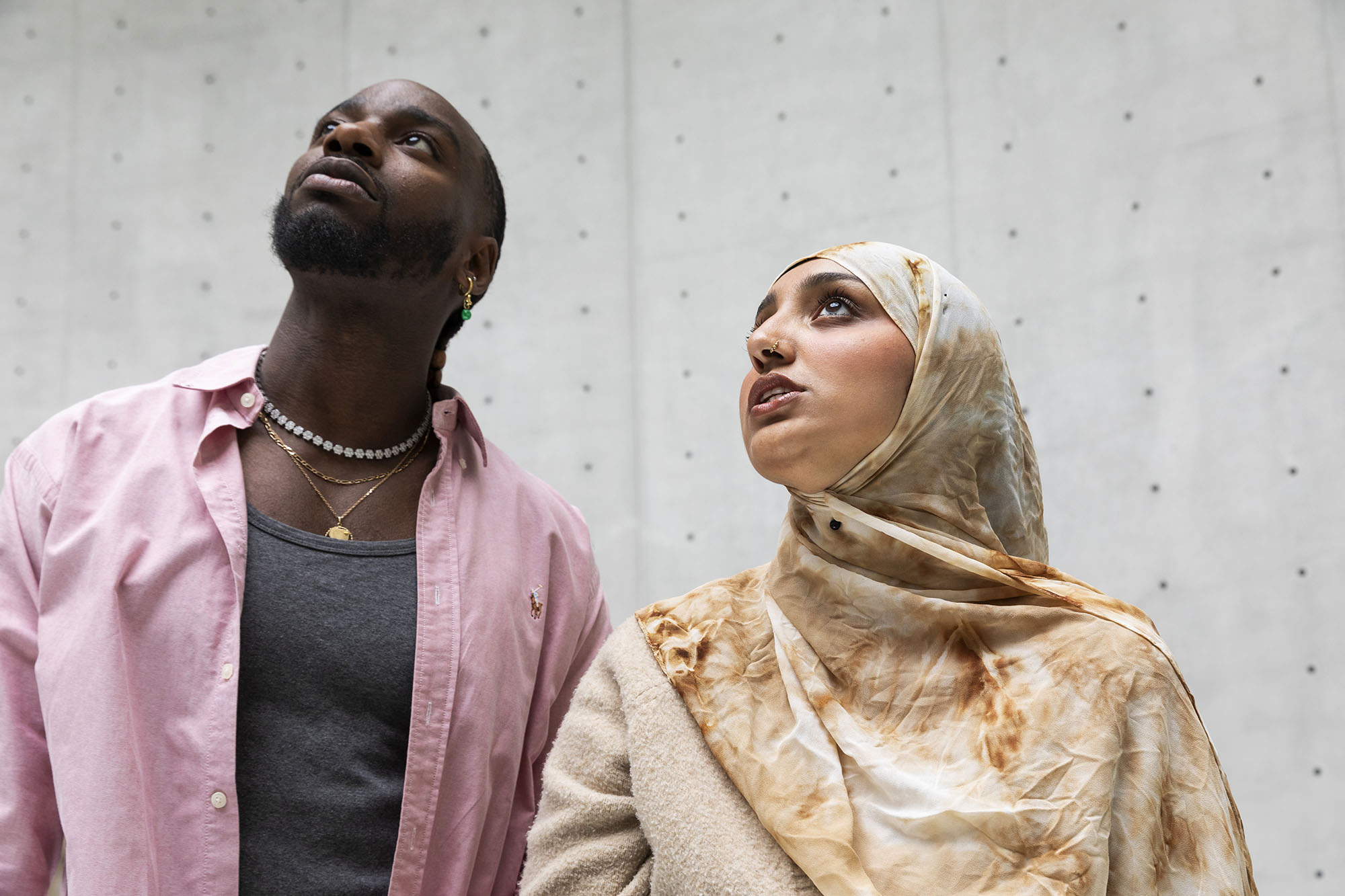 Une femme portant le foulard et un homme en chemise rose regardent vers le haut, derrière eux, un mur en béton gris
