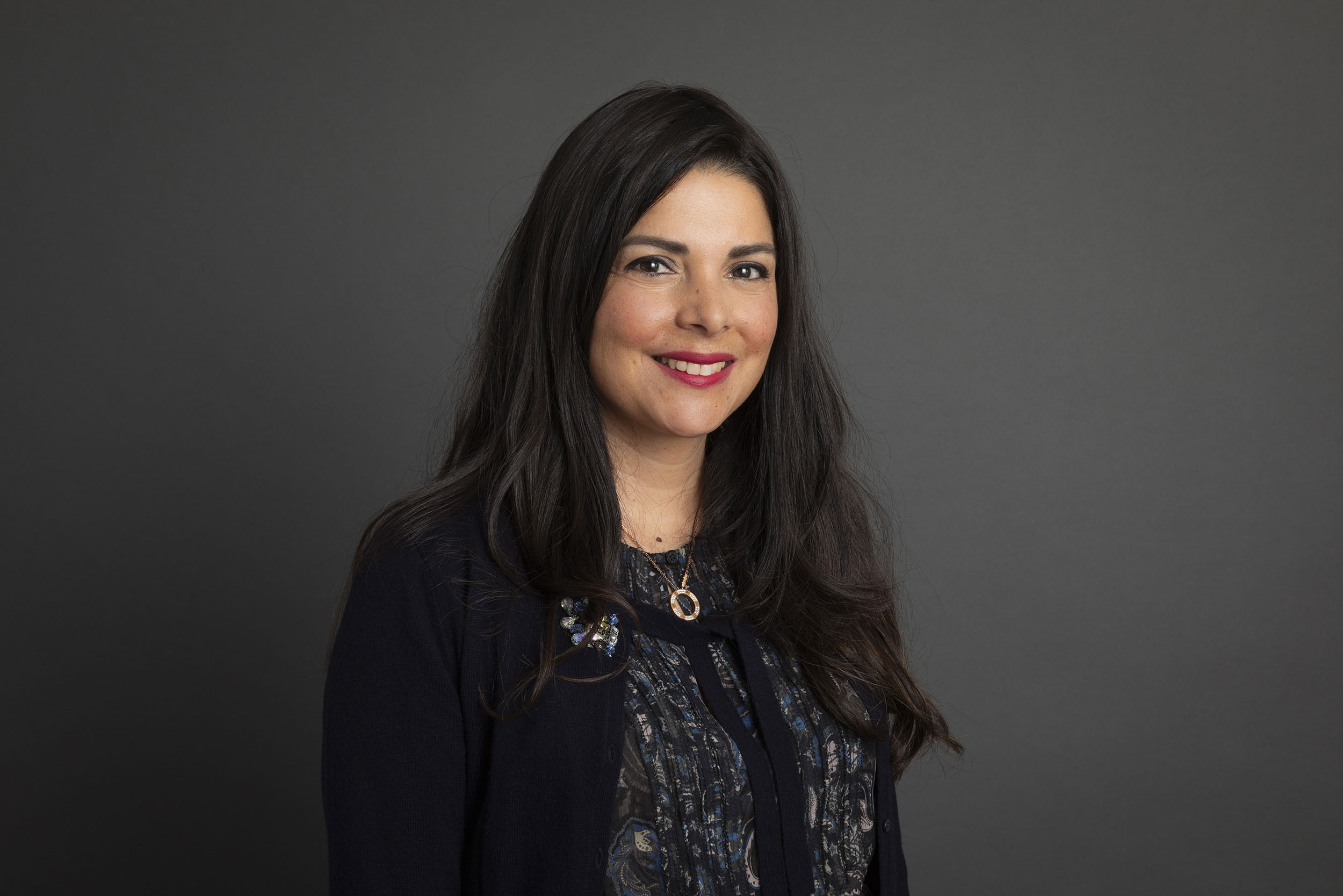 Portrait corporate en studio d'une femme brune souriante aux cheveux long sur fond gris, elle regarde dans l'objectif